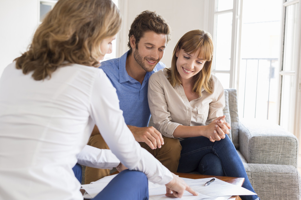 A smiling couple sits on a sofa, attentively listening to a woman pointing at documents on a table. Bright, natural light fills the cozy room.